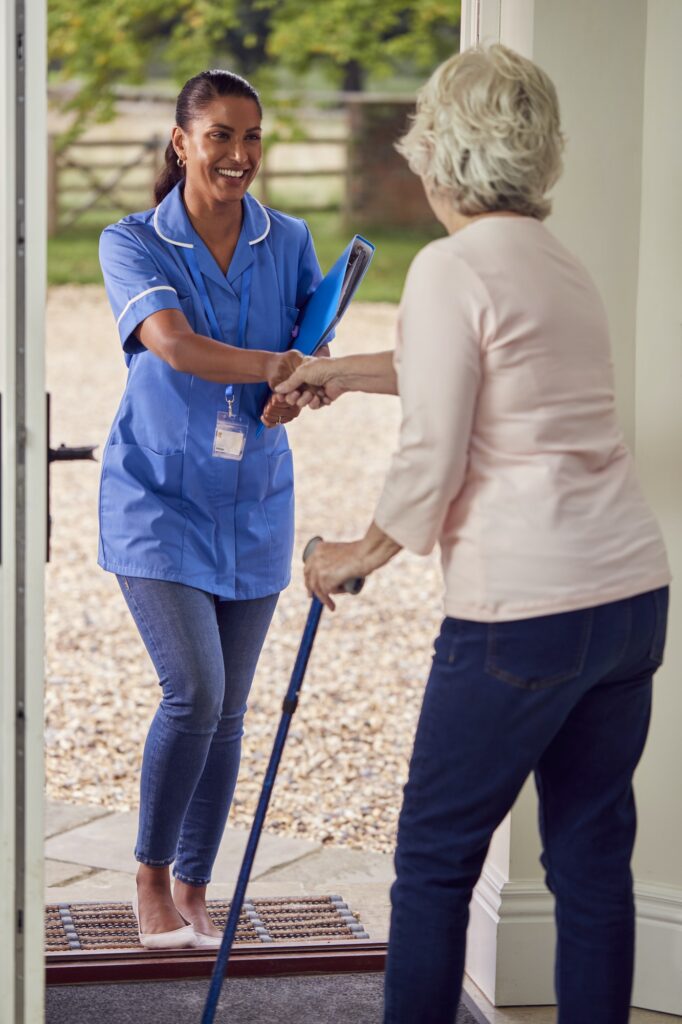 Senior Woman At Home Using Walking Stick Greeting Female Nurse Or Care Worker In Uniform At Door